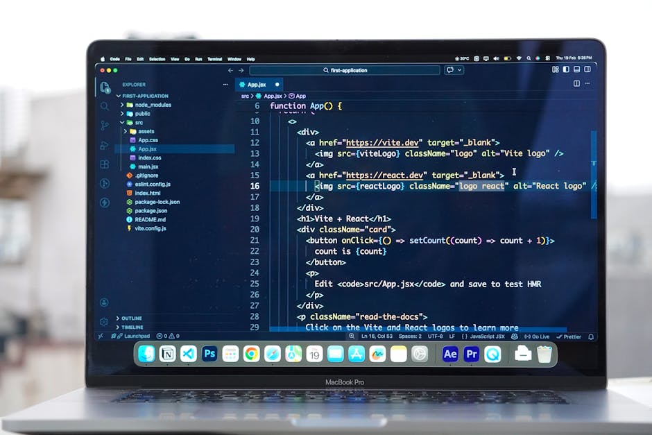 The image displays a close-up of a MacBook Pro laptop on a white desk, with the screen showing a code editor window filled with JavaScript, HTML, and CSS scripts related to a web application. The code